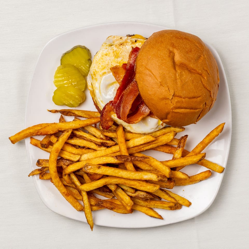A burger with bacon and a fried egg, served with a side of french fries and pickles.