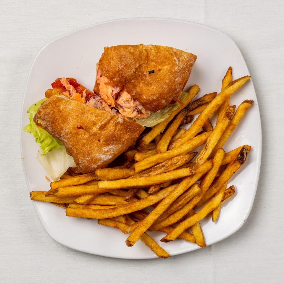 A salmon sandwich on ciabatta bread with lettuce and tomatoes, served with a side of french fries.