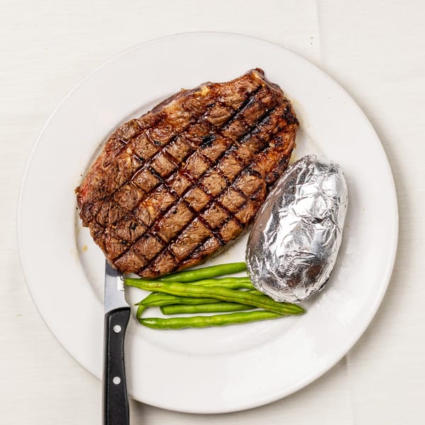 Grilled steak with grill marks, a foil-wrapped baked potato, and green beans on a white plate.